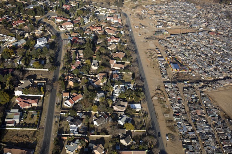 An aerial view shows a mass of shacks on one side and a green, spread out suburb on the other, divided by a wall.