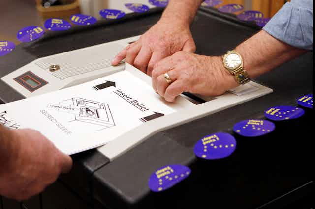 A person's hand feeds a ballot into a scanner while another person's hands help