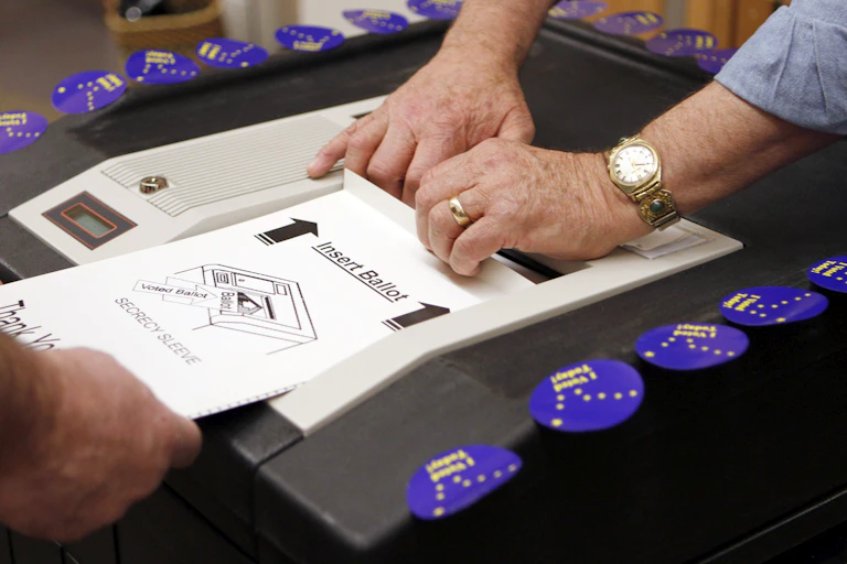 A person's hand feeds a ballot into a scanner while another person's hands help