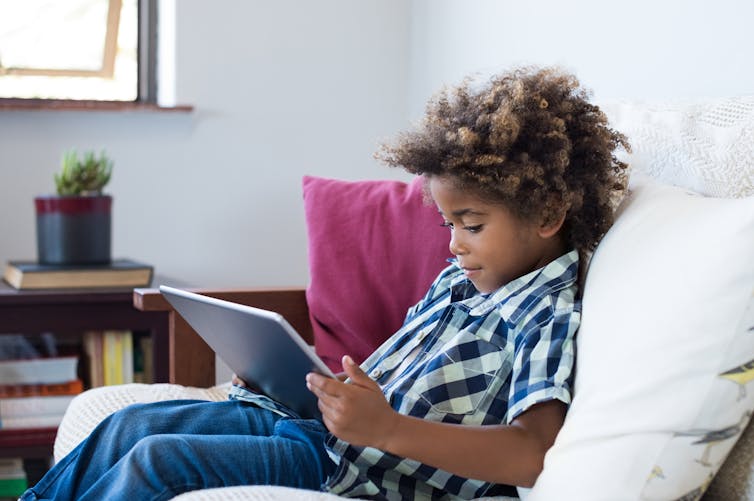 A young black boy using a tablet sits on a sofa