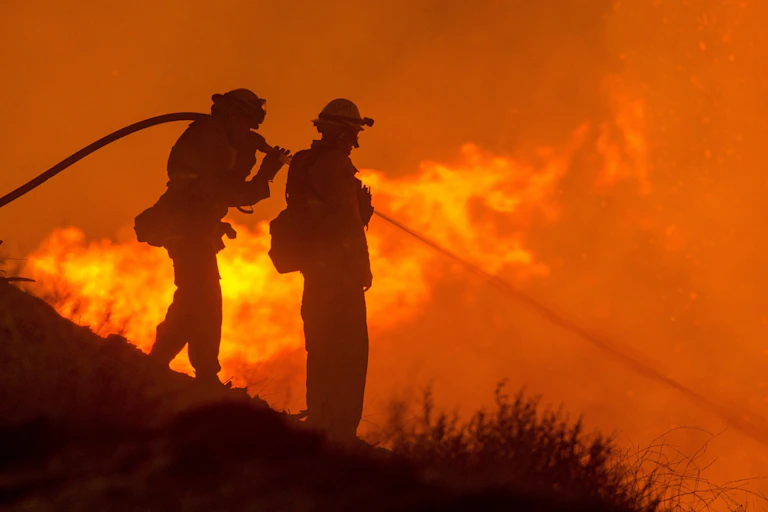 Two firefighters steady a hose on a hillside, spraying water on flames below.