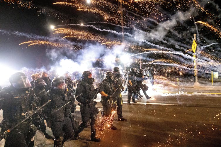 Police in riot gear and carrying weapons walk in a line as the plumes from tear gas cannisters are seen behind them.