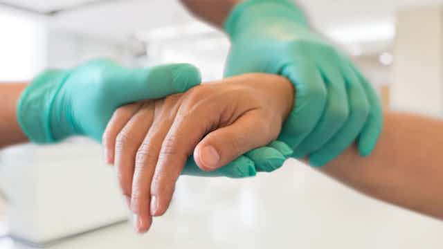 Dos manos con guantes sanitarios sujetan una mano alicaída de persona madura.
