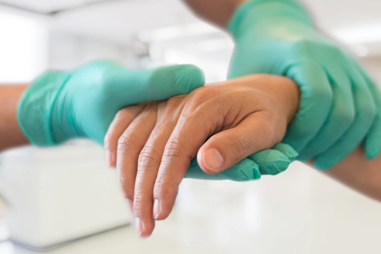 Dos manos con guantes sanitarios sujetan una mano alicaída de persona madura.