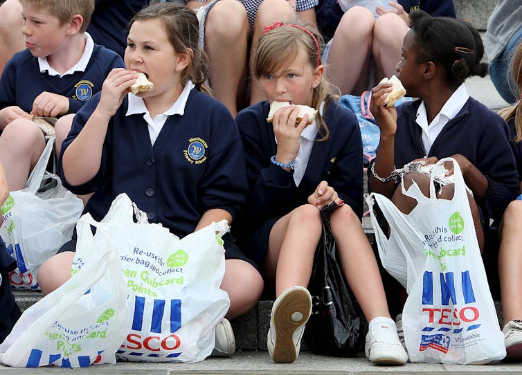 Three children sittiing on the ground eat sandwiches with Tesco bags at their feet.