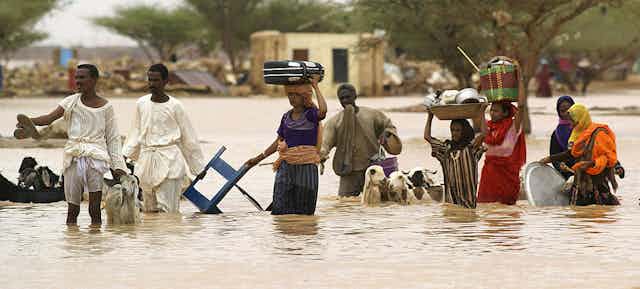 People carrying belongings on their heads and leading goats walk through flooded area
