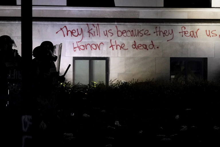 Two police officers in riot gear are seen in silhouette in front of a building with the spraypainted phrase: They kill us because they fear us.