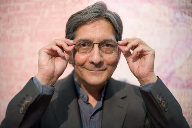 A man in suit jacket grey shirt smiles playfully as he looks into camera holding the sides of his spectacles.