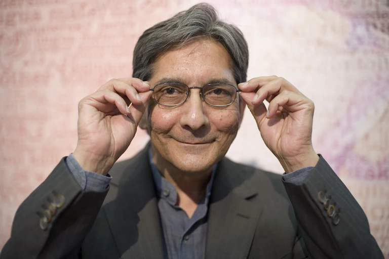 A man in suit jacket grey shirt smiles playfully as he looks into camera holding the sides of his spectacles.
