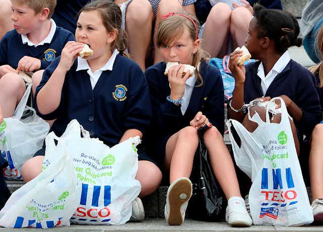 Three children sittiing on the ground eat sandwiches with Tesco bags at their feet.