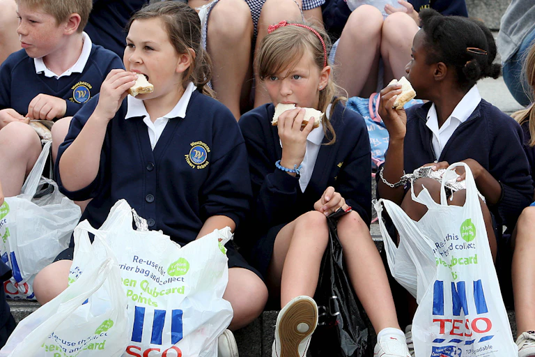 Three children sittiing on the ground eat sandwiches with Tesco bags at their feet.