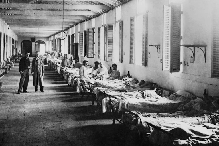 Two men standing next to row of men in hospital ward.