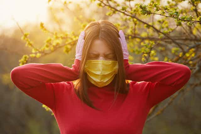 Una mujer con mascarilla amarilla y guantes sanitarios rosas se tapa los oídos con gesto de no desear oír nada.