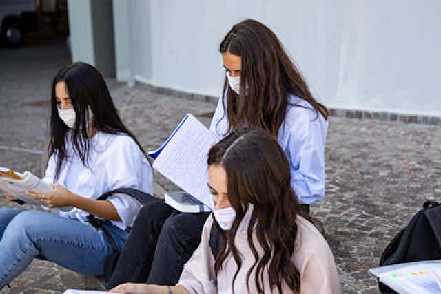 A group of female students sit outside reading notebooks as they wear masks. 