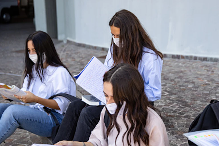 A group of female students sit outside reading notebooks as they wear masks.
