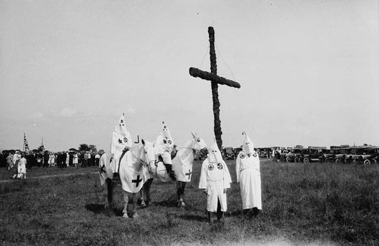 Hooded figures stand in front of a cross