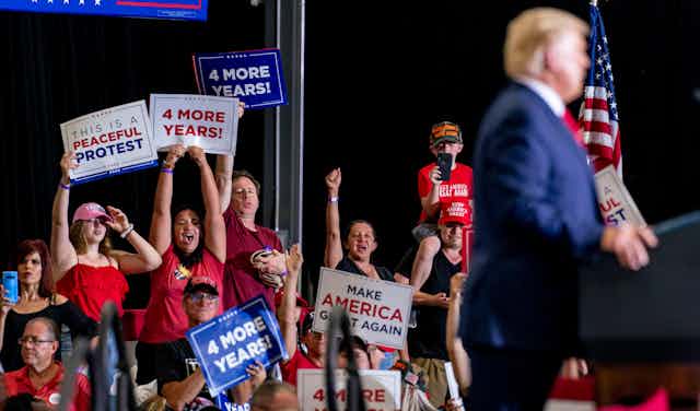 Trump supporters wave signs reading Four More Years and Make America Great Again as Donald Trump speaks behind a podium.