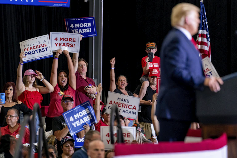 Trump supporters wave signs reading Four More Years and Make America Great Again as Donald Trump speaks behind a podium.