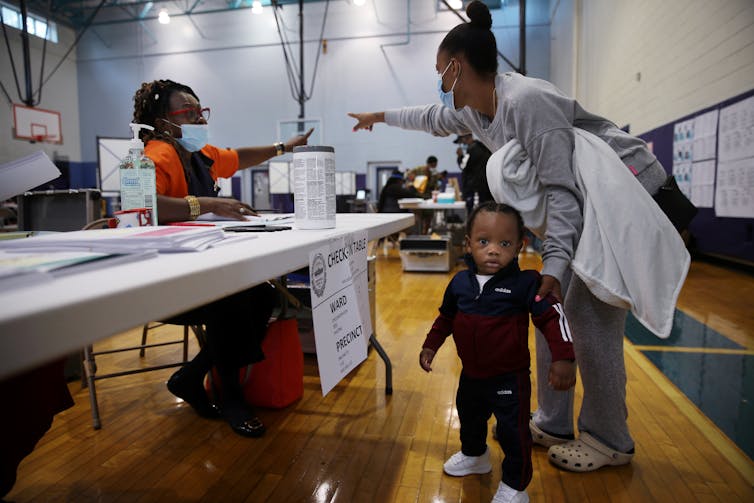 A Black woman and her small son receive instructions from a Black poll worker in a gym set up for voting