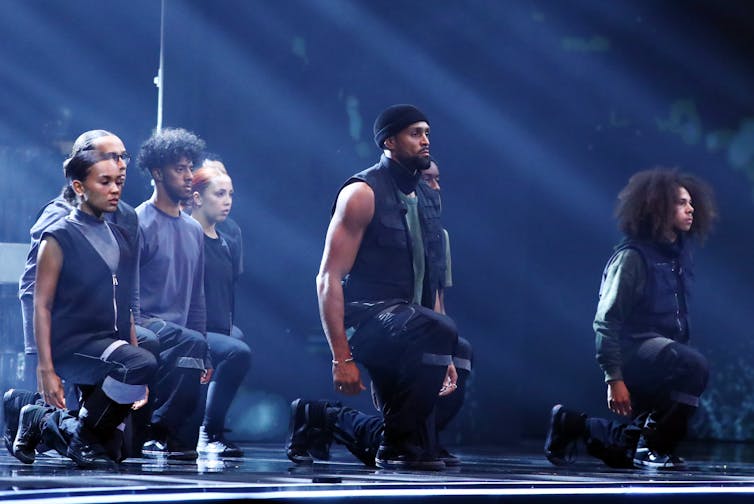 Group of dancers in black kneeling on one knee with their eyes closed.