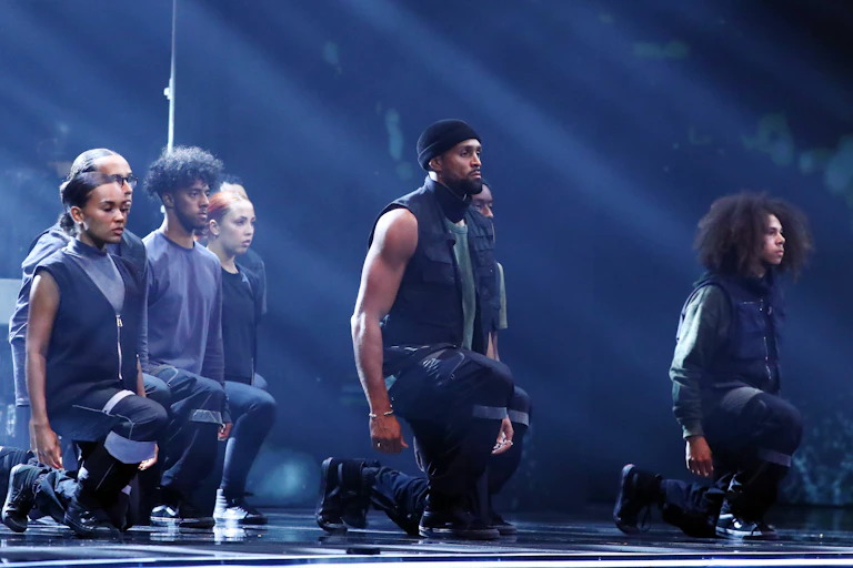 Group of dancers in black kneeling on one knee with their eyes closed.