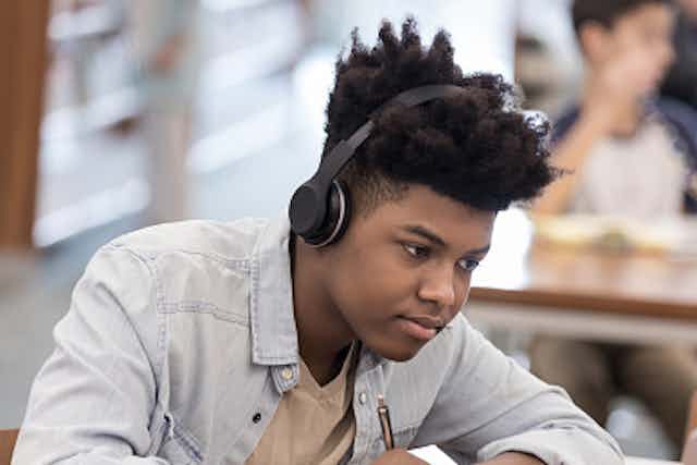 A young African American boy writes on a notebook while reading on his laptop with a headset on.