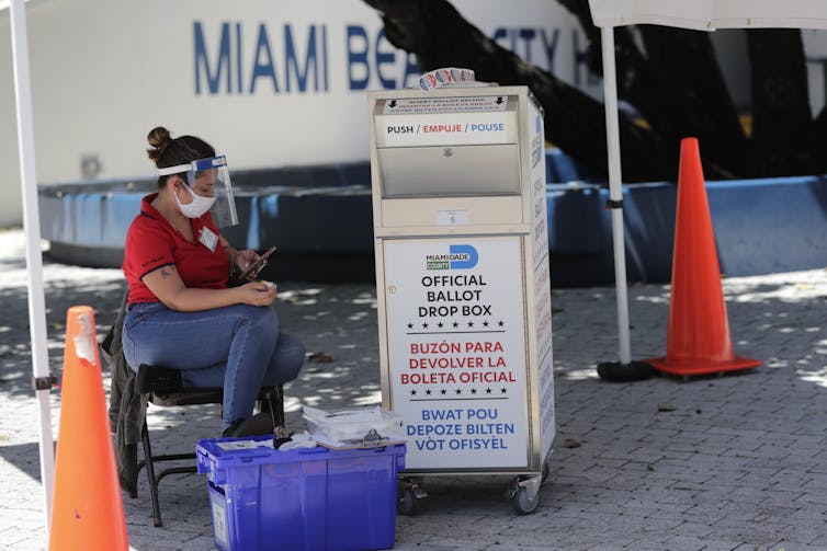 A woman with a mask on sits near an election drop box.