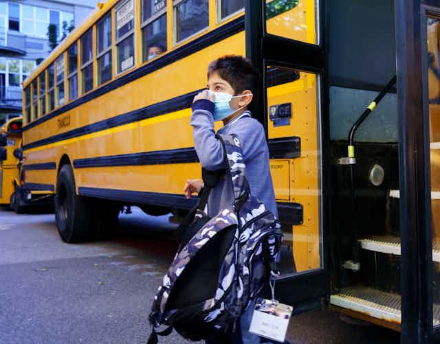A boy stepping off a school bus carry a backpack and adjusting a blue face mask.