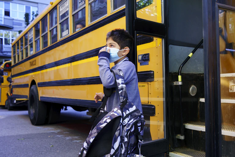 A boy stepping off a school bus carry a backpack and adjusting a blue face mask.