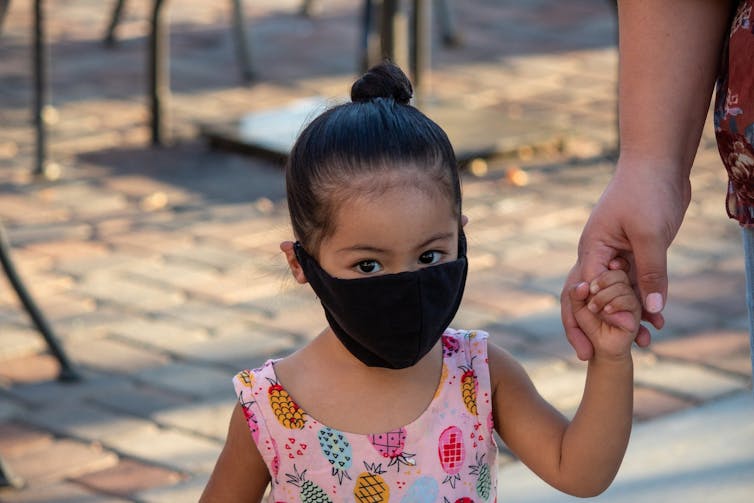 Little girl in a pink dress wearing a black face mask holding the hand of an adult who is out of the frame.