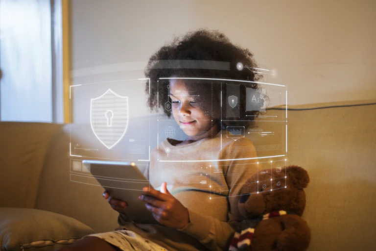 A young girl sitting on a couch, with a teddy bear next to her, looking at the tablet in her hands.