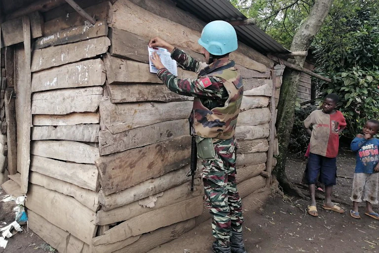 Man in blue helmet pins poster to wall of wooden house with two children looking on.