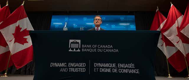 A man sits behind a dais reading Bank of Canada, with Canadian flags on either side.