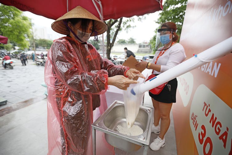 A woman in a pointed hat and rain poncho collects rice from a dispensing machine.