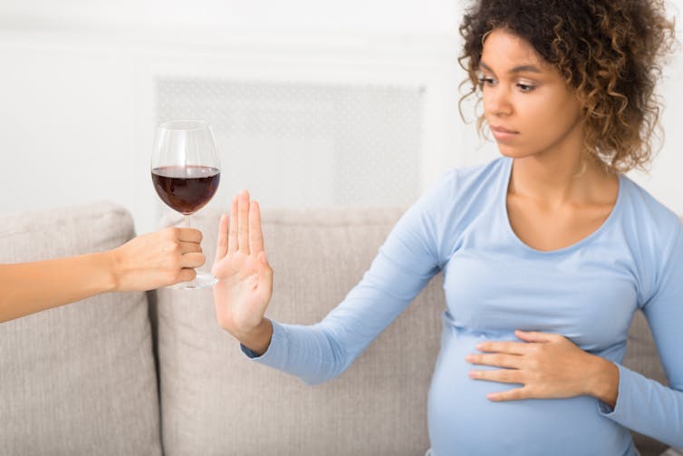 A pregnant woman sitting on a sofa holds up her hand to decline a glass of wine.
