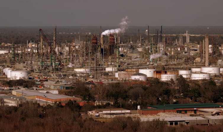 A plume of white gas flowing from a smokestack surrounded by other stacks and oil tanks