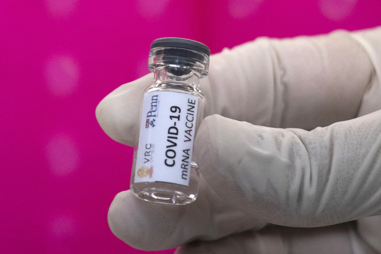 Close-up of gloved fingertips holding a small clear vial labelled "COVID-19 mRNA VACCINE" against a bright pink background