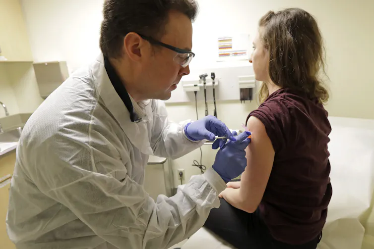 A woman with her back to the camera sits on the examination table in an examination room as a man in a white coat and purple gloves gives her an injection in her upper left arm.