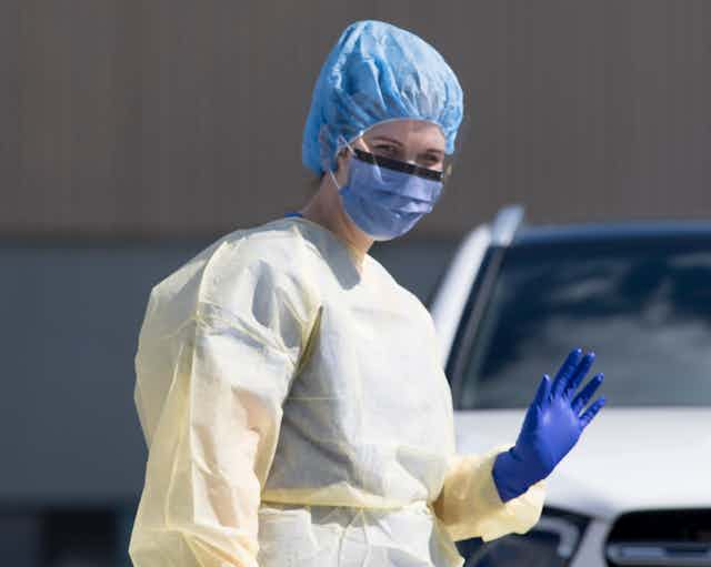 A woman wearing a yellow protective gown, purple gloves and a blue hat and face mask waves her hand.