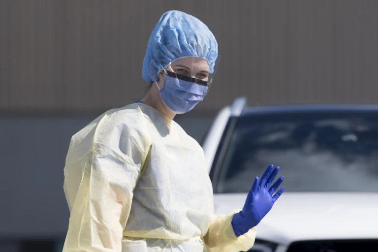A woman wearing a yellow protective gown, purple gloves and a blue hat and face mask waves her hand.