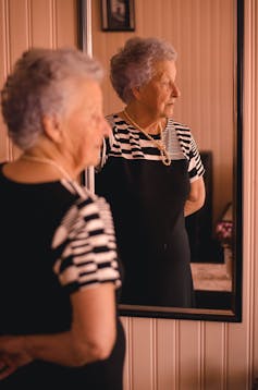 A woman with gray hair in a black dress and pearls stands in front of a mirror, looking off to one side.