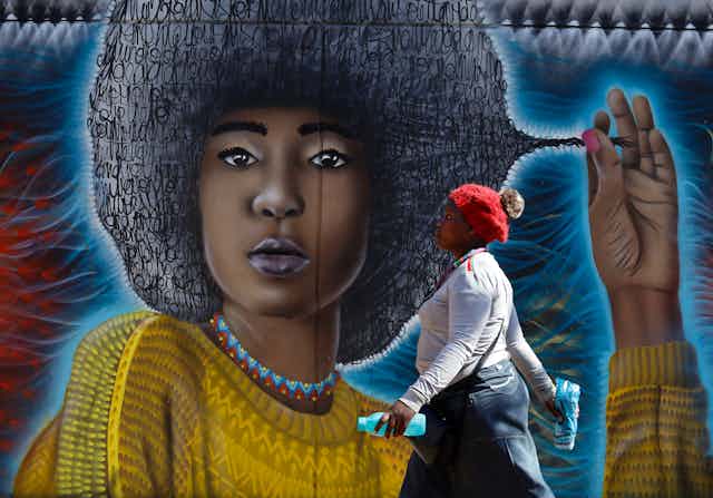 A woman walks past a street art mural showing a young woman with an Afro hairstyle comprised of squiggles, pulling at a lock of it.