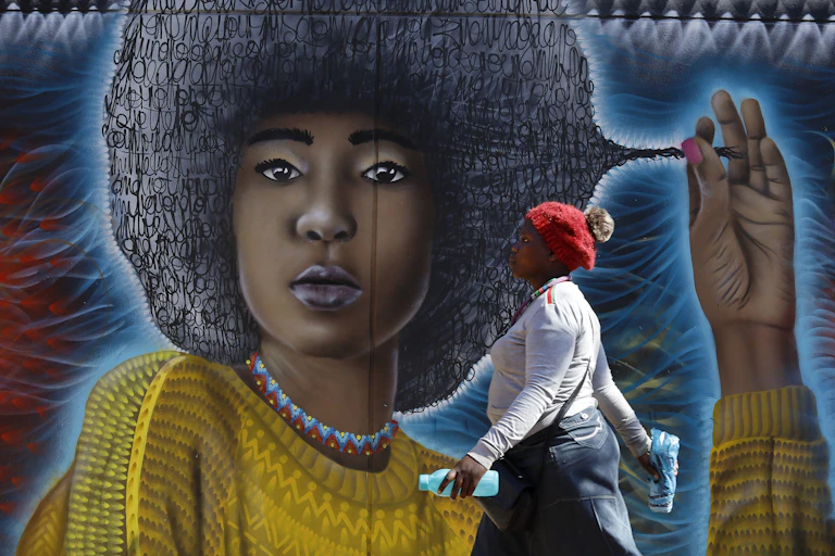 A woman walks past a street art mural showing a young woman with an Afro hairstyle comprised of squiggles, pulling at a lock of it.