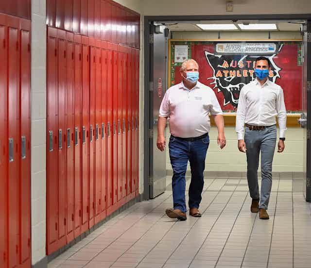 Premier Doug Ford and Education Minister Stephen Lecce, casually dressed in jeans, white shirts and blue face masks, walk beside a row of red lockers. 