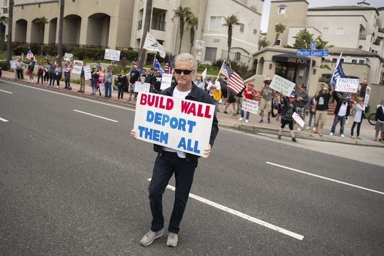 An anti-immigration protester stands in the street holding a sign that reads, 'Build Wall Deport Them All'