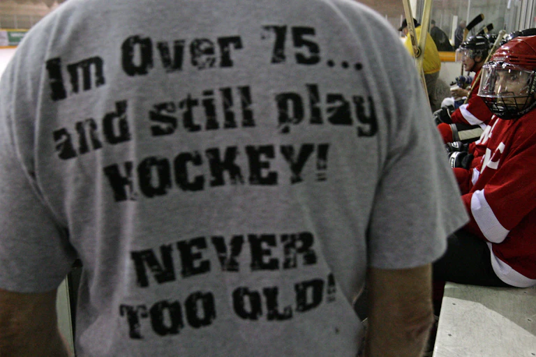 A man at a hockey rink wears a t-shirt that says I'm over 75...and still play HOCKEY! NEVER TOO OLD!