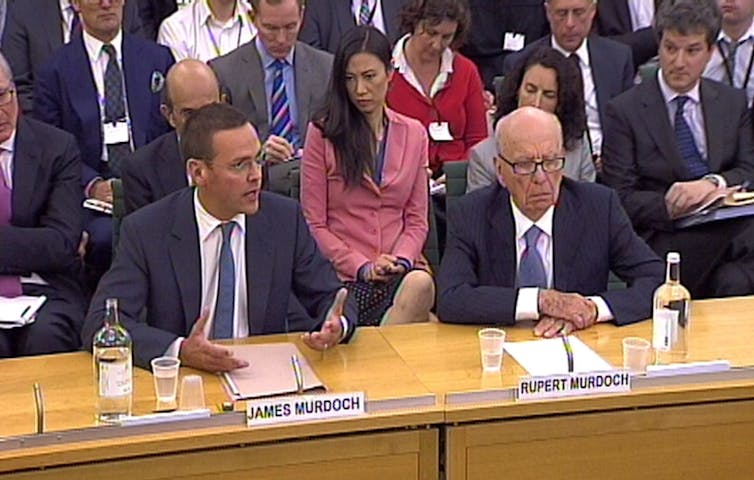 Two men in suits sit behind a wooden desk, giving evidence.