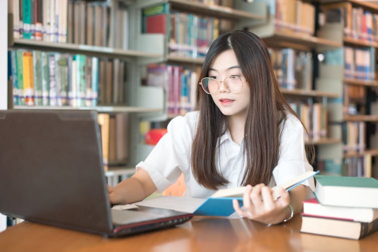 A young woman uses a laptop while holding a book in her hand.
