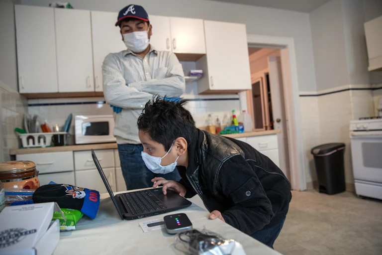 A boy in a mask stares into his laptop while his father looks on.