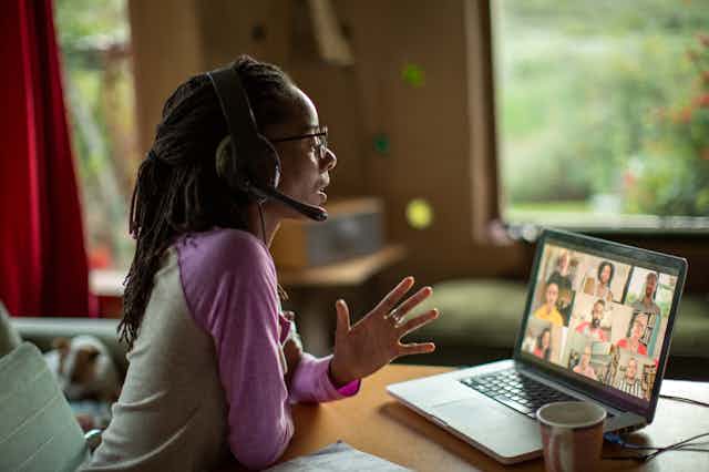 a woman wearing a headset in a home Speaks and gestures in front of an open laptop displaying a video conference